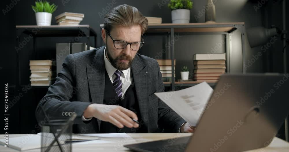 Responsible mature man in trendy suit and eyeglasses sitting at table at own office and working on laptop during evening time. Bearded office manager skilfully doing his job Responsible mature man in trendy suit and eyeglasses sitting at table at own office and working on laptop during evening time. Bearded office manager skilfully doing his job