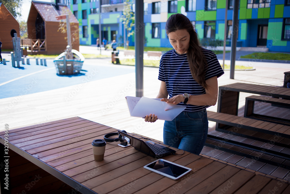 A serious girl is standing in front of a laptop, intently studying the documents and papers she holds in her hands.