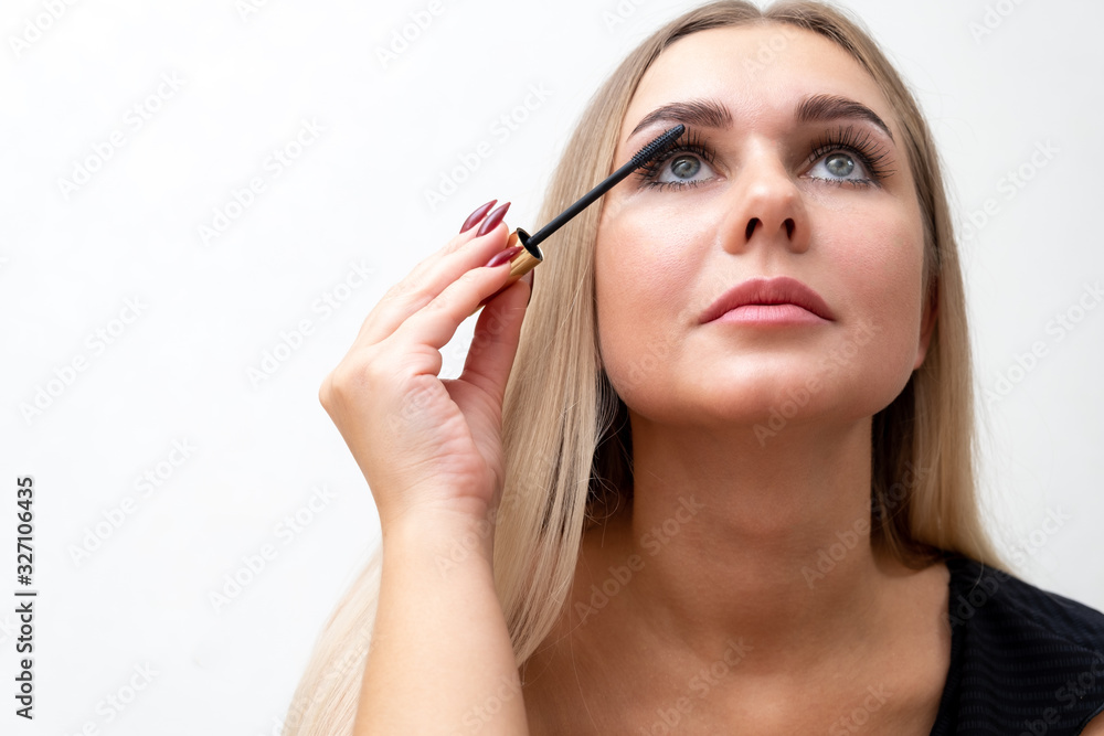 Woman puts black mascara on her eyelashes with makeup, photo of attractive blonde on a white and gray background. Woman puts black mascara on her eyelashes with makeup, photo of attractive blonde on a white and gray background.