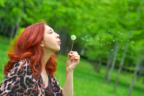 Walk in the Park. Portrait of a beautiful young laughing woman with red hair sitting on meadow with dandelion in hand, blurred background.