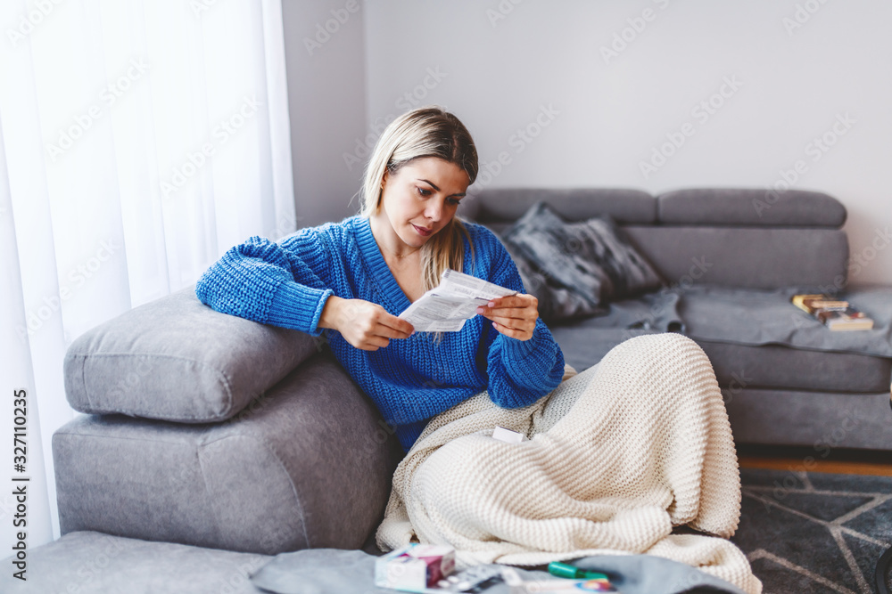 Attractive caucasian blond woman sitting on sofa in living room covered with blanket and reading side effects about new medicine. - obrazy, fototapety, plakaty 
