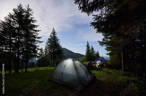Evening camping in the mountains. Friends are sitting near tents in which the light is on between the fir trees under the evening sky on which stars with a magical landscape of mountains appear.