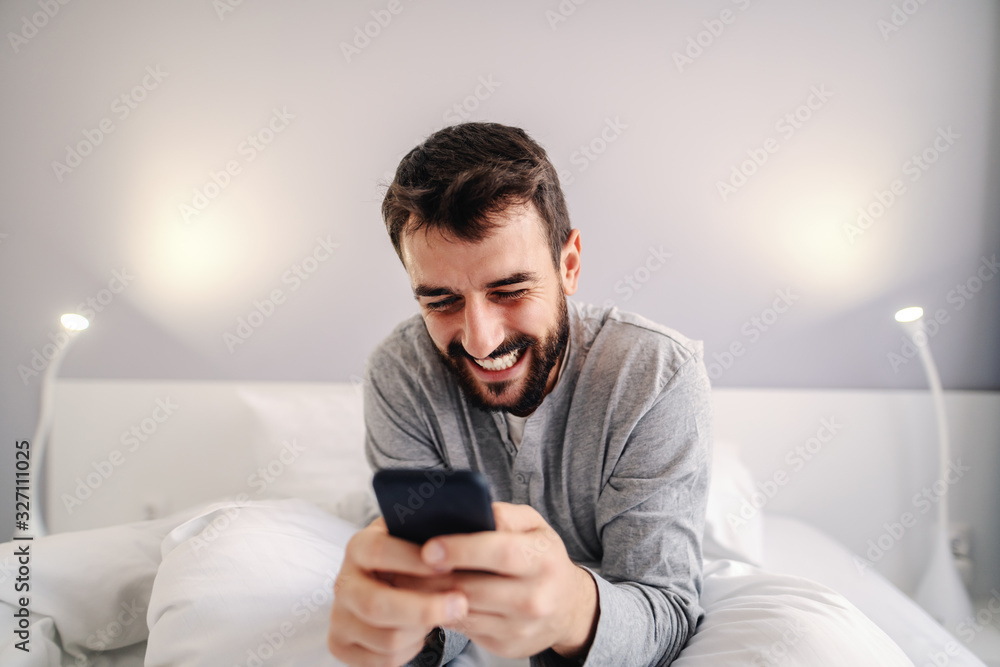 Young attractive smiling bearded caucasian man sitting on bed in bedroom and using smart phone for late night online chatting with his girlfriend. - obrazy, fototapety, plakaty 