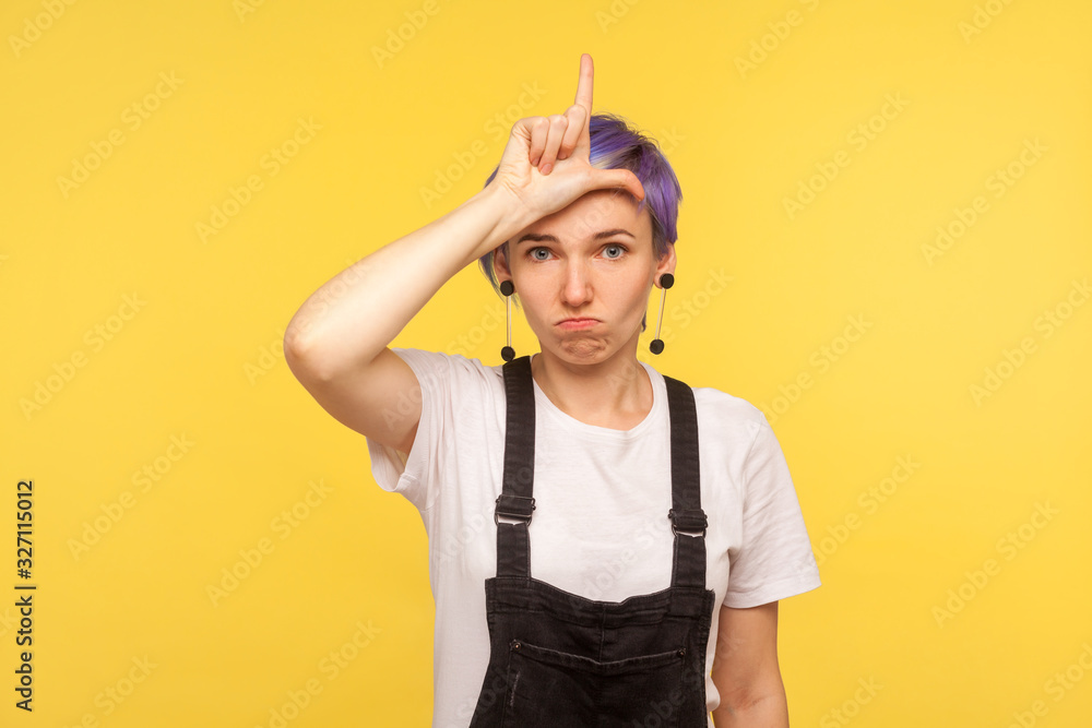 Loser! Portrait of displeased upset hipster girl with violet short hair in denim overalls showing loser gesture, L finger symbol on head, worried about failure, mistake. yellow background, studio shot Loser! Portrait of displeased upset hipster girl with violet short hair in denim overalls showing loser gesture, L finger symbol on head, worried about failure, mistake. yellow background, studio shot
