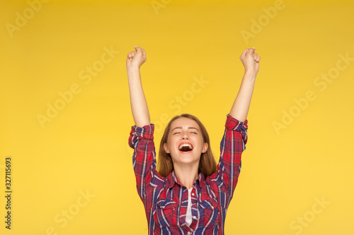 Hurray, victory! Portrait of delighted girl in checkered shirt yelling for happiness, enjoying life, rejoicing success with arms raised, celebrating triumph. studio shot isolated on yellow background