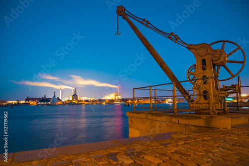 Old port crane with the working factory background at night in Copenhagen, Denmark. Larsens Plads Amaliekaj famous quay near Nyhavn. long exposure lights & clear sky with stars. Old European buildings