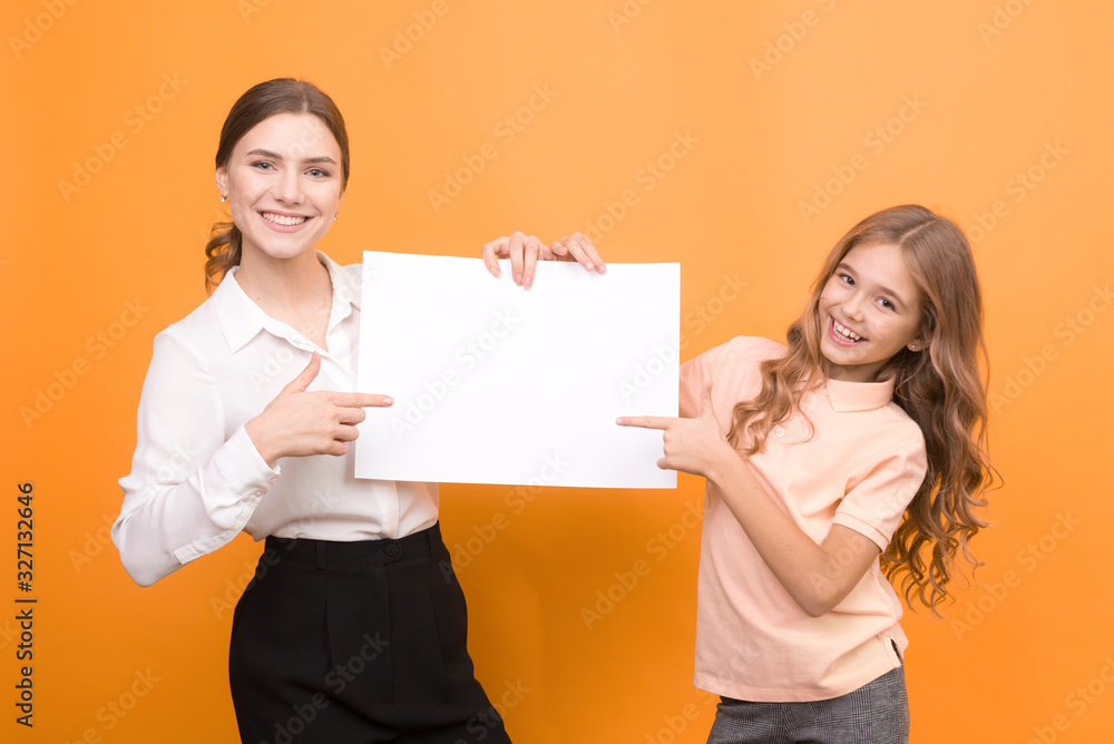 two funny girls holding clear sheet of paper on orange background. mock up. two funny girls holding clear sheet of paper on orange background. mock up.