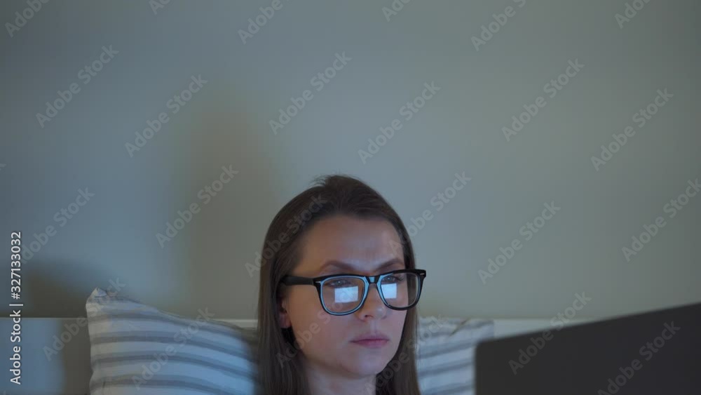 Woman in glasses working on laptop while lying in bed at night. Mobile addict or insomnia concept. Woman in glasses working on laptop while lying in bed at night. Mobile addict or insomnia concept.