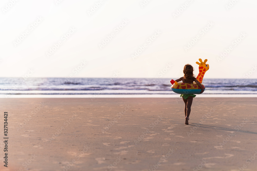 A happy girl with a rubber giraffe ring running across the beach to the sea. Back view. Sunset on a beach. Vacation and summer concept. Happy childhood concept.