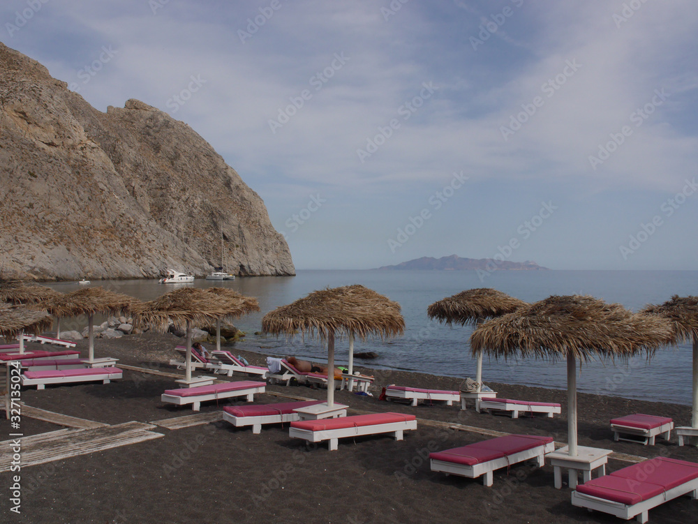 Parasols and empty sun loungers on the black sand of Perissa Beach. Parasols and empty sun loungers on the black sand of Perissa Beach.