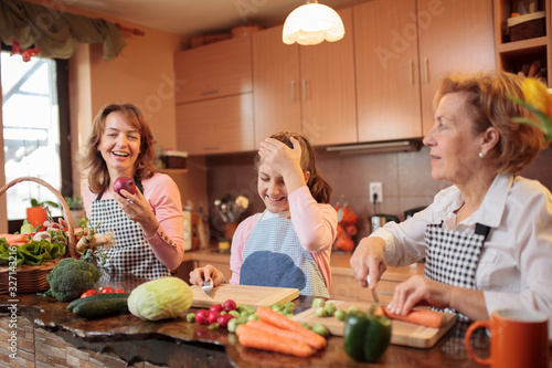 Young caucasian teenage girl preparing food, helping her mother and grandmother by cutting fresh vegetables in home kitchen photo