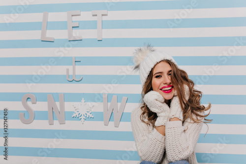 friendly, kind young girl with beautiful bright lipstick smiling sweetly, touching her face with her hands in mittens. Portrait of posing smiling model with curls sitting against striped blue wall