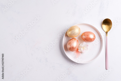 Greeting card with handmade painted bright eggs on a plate and golden spoon on a light grey marble background.
