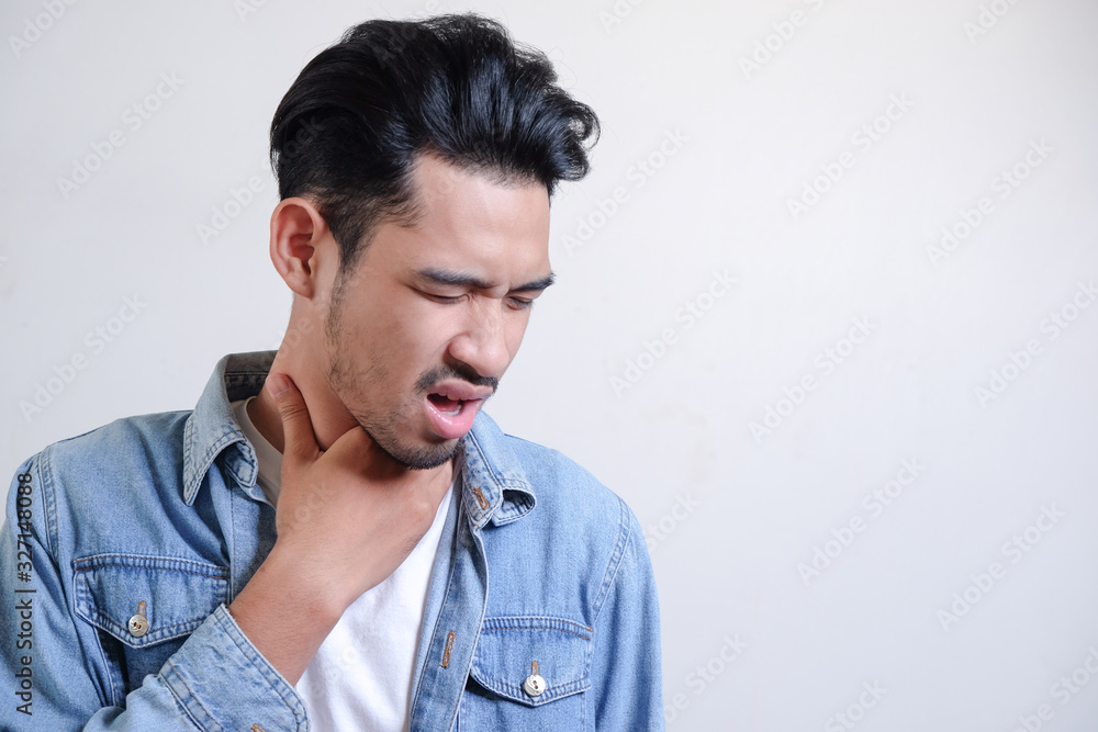 Fototapeta premium Young man have a sore throat and touching his neck, wearing white t-shirt and blue jeans jacket against light grey background, hard to swallow, wuhan virus infection and public widespread