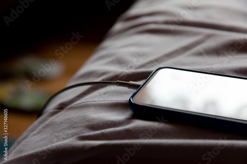 A simple modern no name smartphone laying in bed in crumpled white sheets charging in the morning, closeup. Bedroom background. Leaving a phone device unattended, charging overnight abstract concept photo