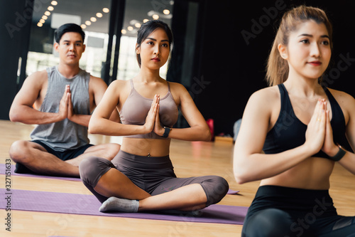 Group of Asian women and man doing pilates lying on yoga mats in aerobics class. Young sporty people on crossed legs on the floor and do namaste pose in gym studio.