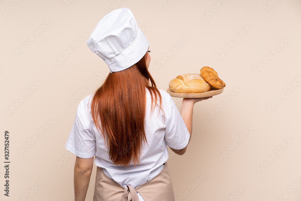 Redhead teenager girl in chef uniform. Female baker holding a table with several breads in back position Redhead teenager girl in chef uniform. Female baker holding a table with several breads in back position