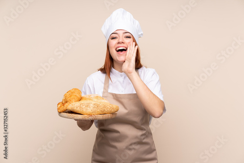 Redhead teenager girl in chef uniform. Female baker holding a table with several breads shouting with mouth wide open