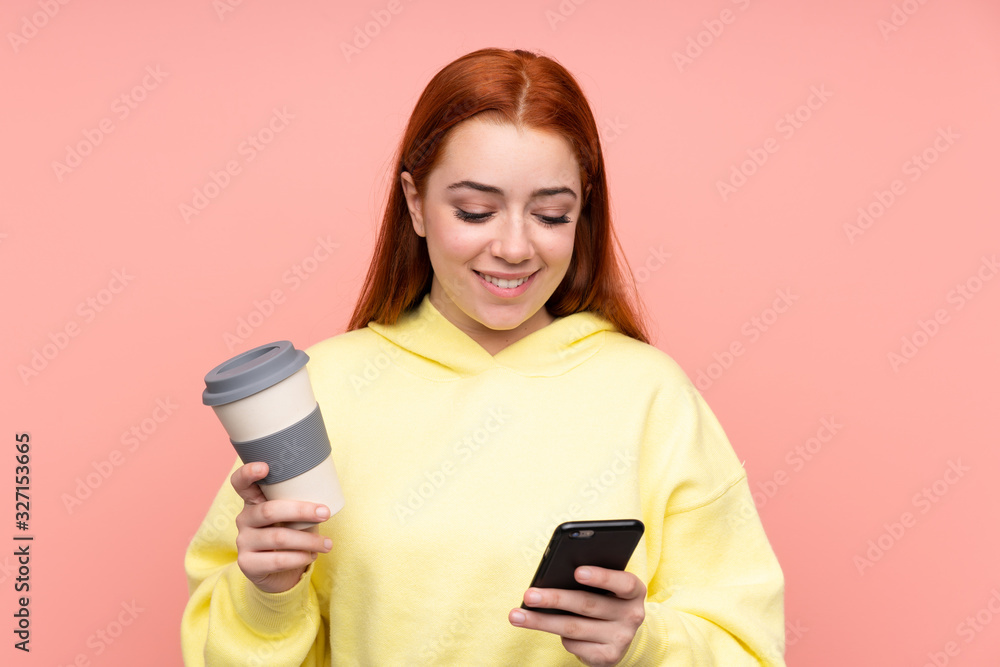 Redhead teenager girl over isolated pink background holding coffee to take away and a mobile Redhead teenager girl over isolated pink background holding coffee to take away and a mobile