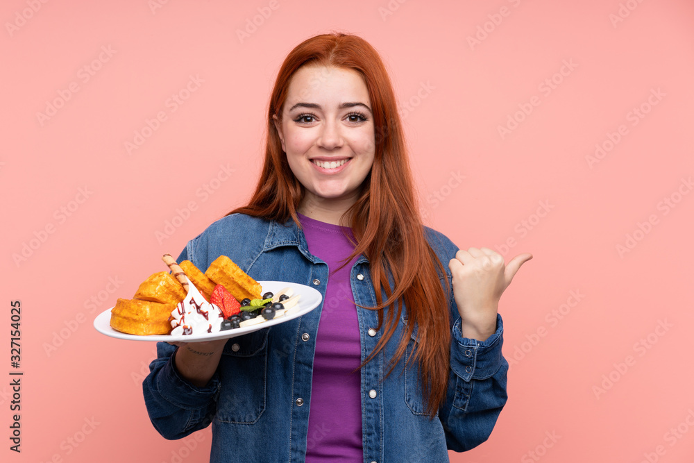 Redhead teenager girl holding waffles over isolated pink background pointing to the side to present a product