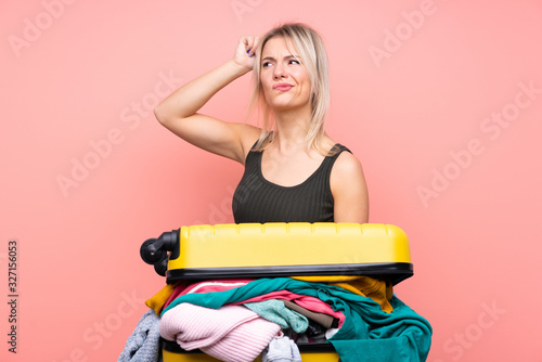 Traveler woman with a suitcase full of clothes over isolated pink background having doubts and with confuse face expression photo