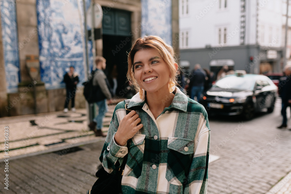 Fototapeta premium Close-up portrait of cheerful white woman with backpack on blur background. Photo of fashionable girl with beautiful brown hair smiling to camera.