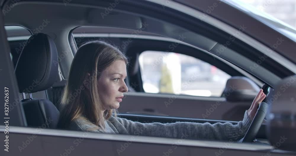 Young confident Caucasian woman sitting in automobile and touching steering wheel. Lady choosing vehicle in car dealership. Side view, close-up. Cinema 4k ProRes HQ.