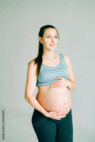 a young pregnant girl in sportswear is standing on a yoga Mat, smiling and holding her belly in the Studio