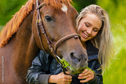 Smiling beautiful woman feeding her arabian horse with grass in the field