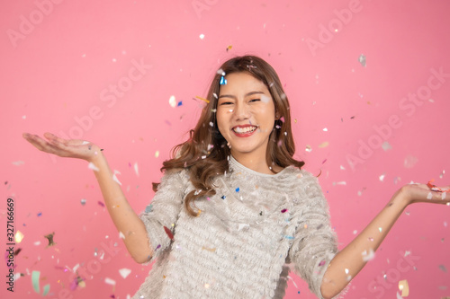 Portrait of a cheerful beautiful Asian womanl wearing dress standing standing under confetti rain and celebrating isolated over pink background