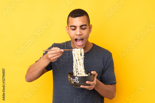 Valokuvatapetti Young African American man over isolated yellow background holding a bowl of noo