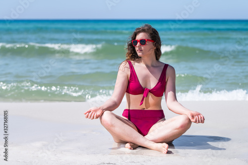 Young girl woman meditating yoga pose in bikini bathing suit by ocean sea green water in background in Santa Rosa Beach and horizon