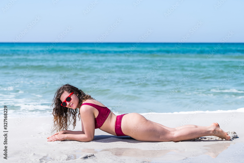 Young happy girl woman lying down on sand in red swimsuit bikini bathing suit sunglasses smiling with ocean shore background in Florida Young happy girl woman lying down on sand in red swimsuit bikini bathing suit sunglasses smiling with ocean shore background in Florida
