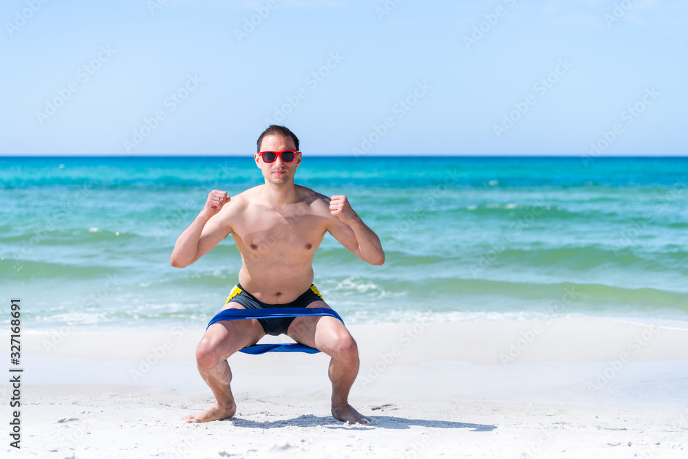 Young adult man squatting at beach working out with elastic resistance band for legs exercise with ocean background Young adult man squatting at beach working out with elastic resistance band for legs exercise with ocean background