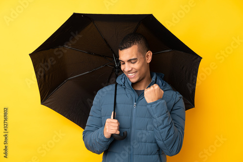 Young handsome man holding an umbrella over isolated yellow background celebrating a victory