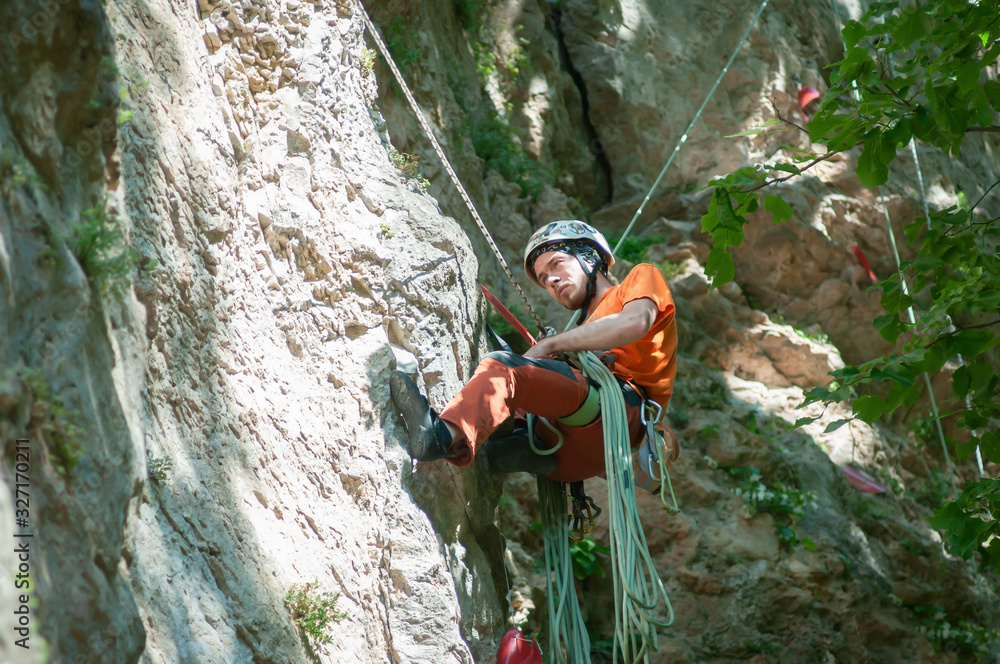 A climber in a helmet and orange t-shirt, with a length of rope and climbing gear with a serious face hanging on a rope on a cliff.