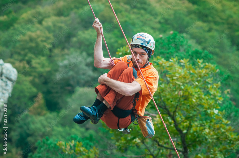 A funny, naughty climber in an orange shirt swinging on a rope on the background of green forest with tucked up knees.
