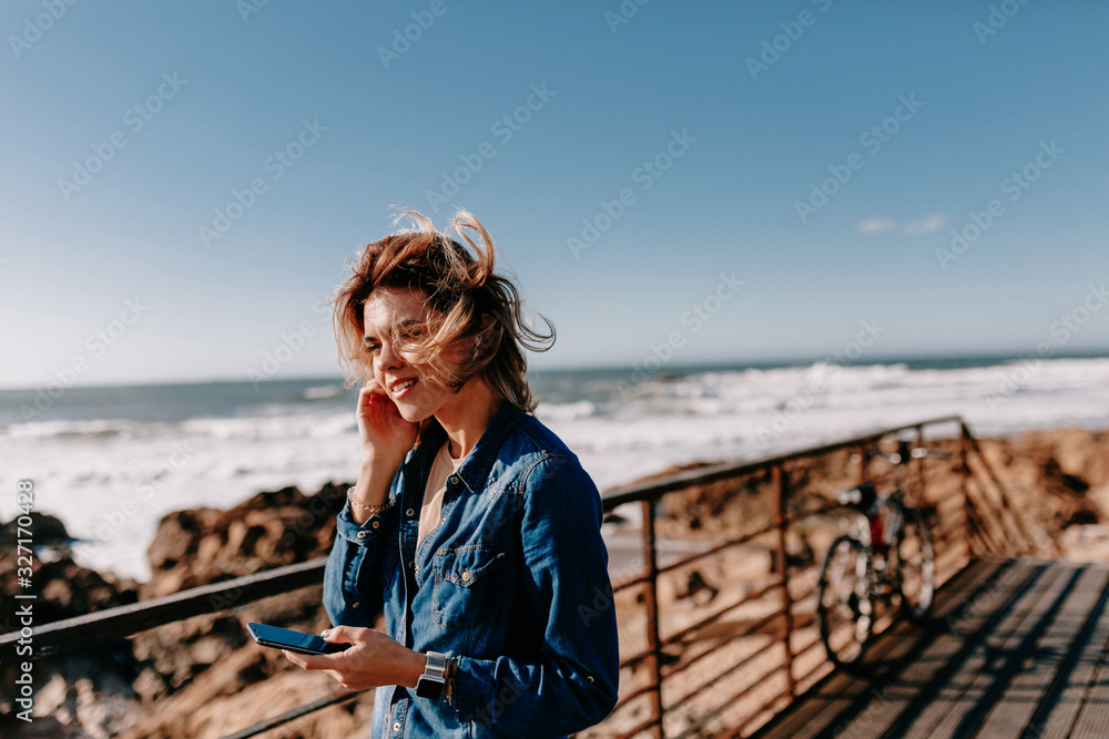 European stylish young woman with flying hair listening music and relaxing near the ocean in sunny day