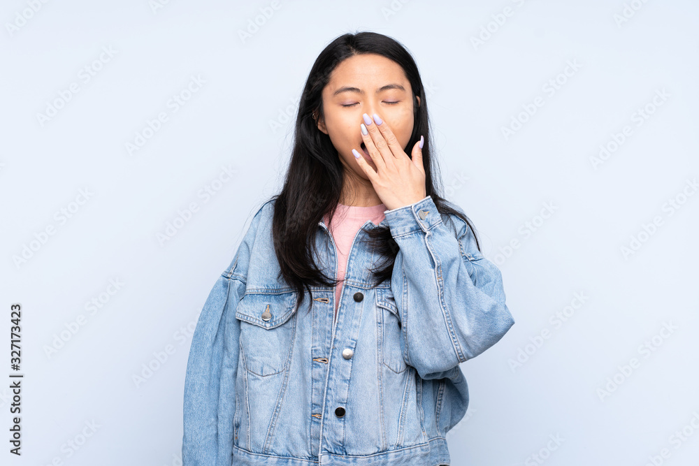 Teenager Chinese woman isolated on blue background yawning and covering wide open mouth with hand Teenager Chinese woman isolated on blue background yawning and covering wide open mouth with hand