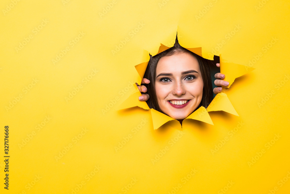 Glad young man with toothy smile looks positively aside, shows face in paper hole, isolated over yellow background with blank space. Positive emotions. Man peeks through ripped paper