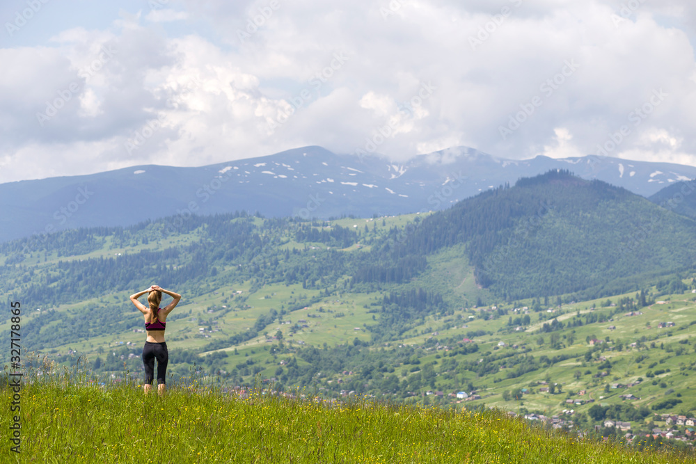 Slim young woman with raised arms outdoors on background of beautiful mountain landscape on sunny summer day.