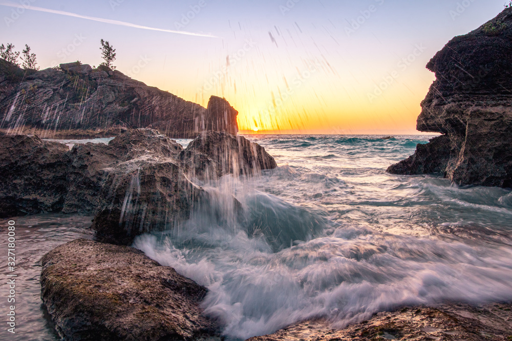 Obraz premium Water droplets spray from a large wave crashing on the rocky coast. Sun just above the horizon in the background - Jobsons Cover Bermuda