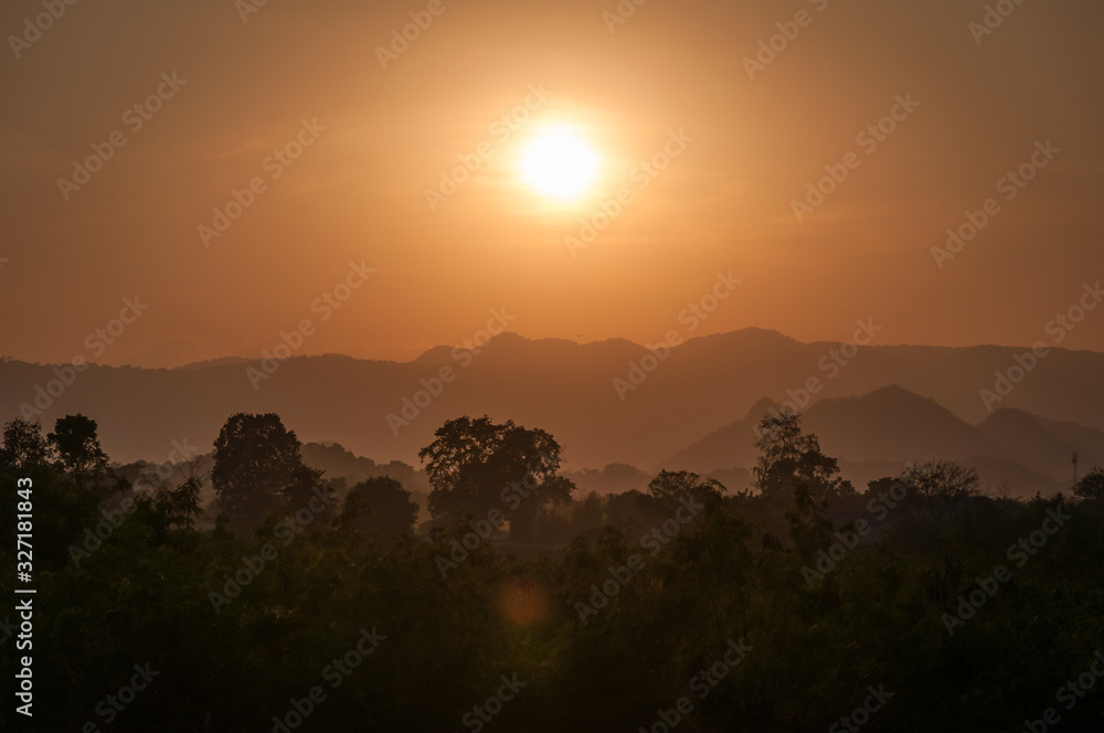 silhouettes of trees and layered mountains in an orange light with a setting sun in the background