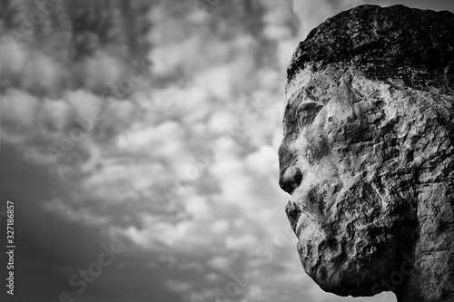 Black and white photo of a sculpture head close up against a cloudy sky.
