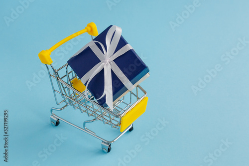 Gift with a bow in a yellow shopping basket on a light blue background. Selective focus.