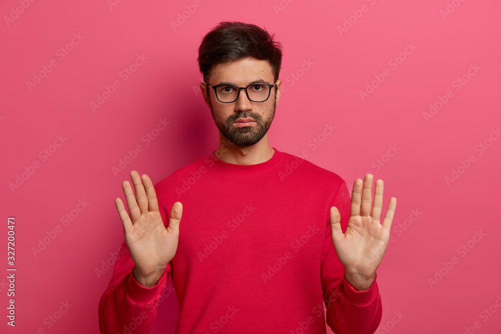 Naklejka premium Photo of European man with thick stubble makes stop gesture, hold sign, pulls palms towards camera, looks seriously, has determined expression, asks to calm down, wears red sweater, glasses.