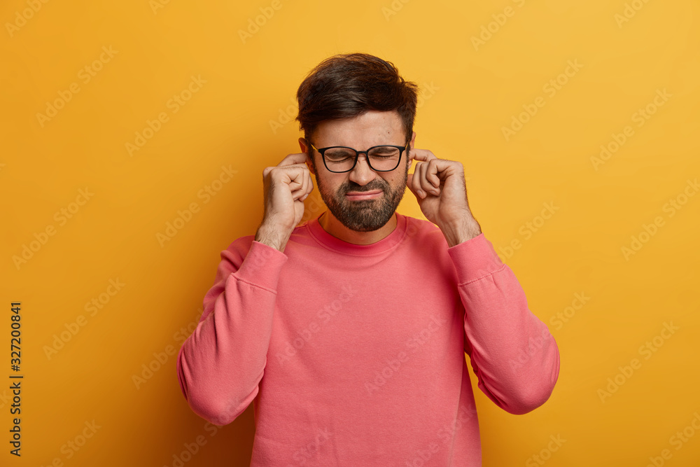 Disturbed bothered man plugs fingers at ear holes, hears annoying loud sound, noise or siren, frowns face, dressed casually, poses over yellow wall, ignores heartbreaking scream coming from other room Disturbed bothered man plugs fingers at ear holes, hears annoying loud sound, noise or siren, frowns face, dressed casually, poses over yellow wall, ignores heartbreaking scream coming from other room