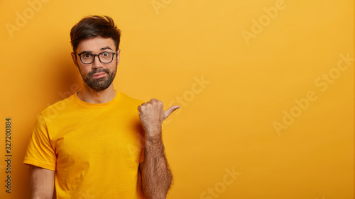 Photo of wondered bearded man points thumb right on copy space, shows amazing sale discount, demonstrates shopping offer, wears glasses and yellow casual t shirt, has worried expression, poses indoor