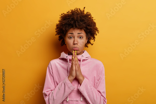 Fototapeta Photo of dissatisfied Afro American woman keeps palms pressed together, prays or