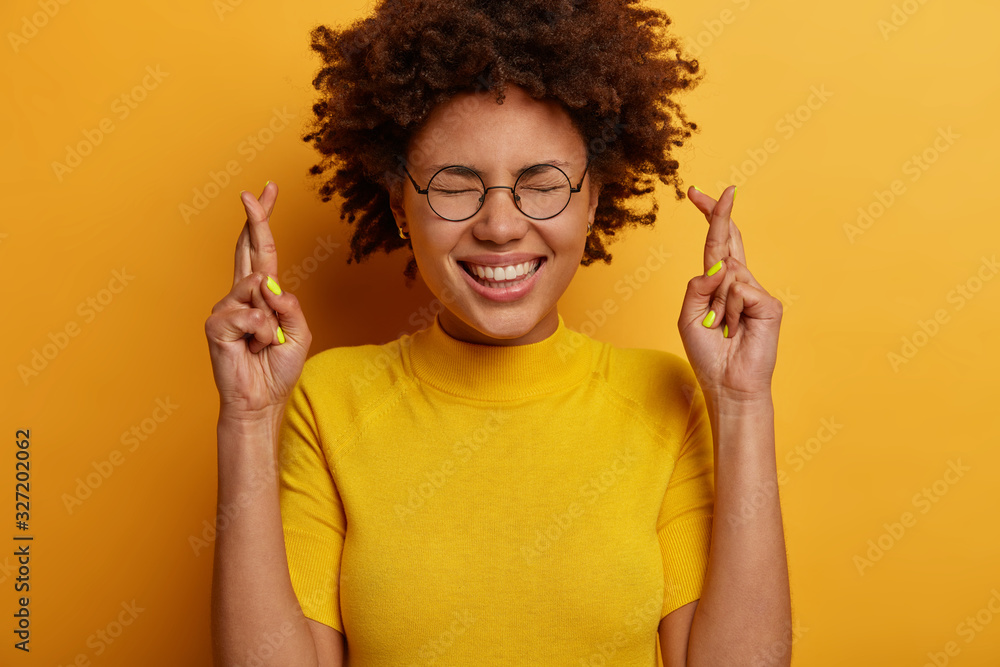 Φωτογραφία Close up shot of cheerful ethnic girl keeps faith, crosses fingers, beggs for go